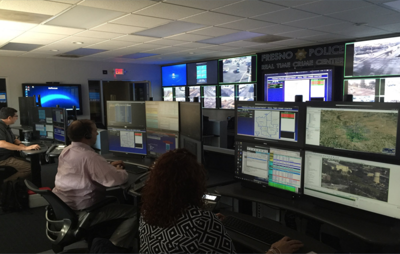 Police sit at workstations in front of a wall of video monitors. 